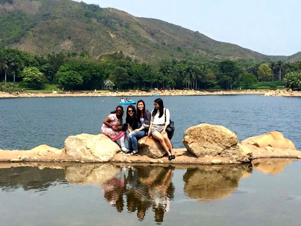 Four people sit together on rocks at the edge of a lake, their reflection in the still water; a wooded hill rises behind.