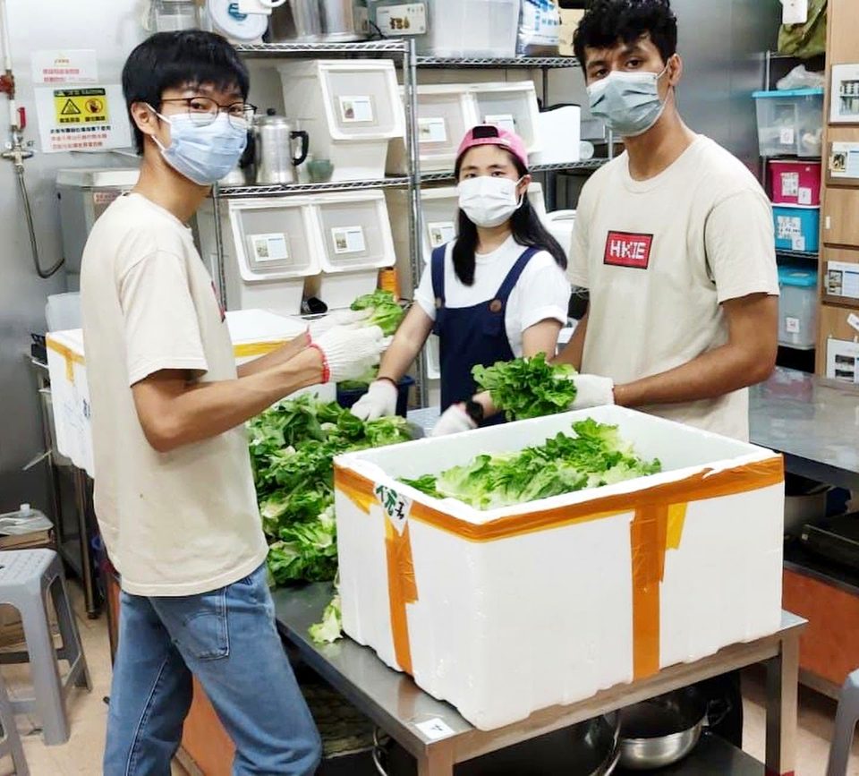 Three people in face masks and work gloves sort leafy vegetables into white foam boxes in a kitchen; one wears a T-shirt printed HKIE.